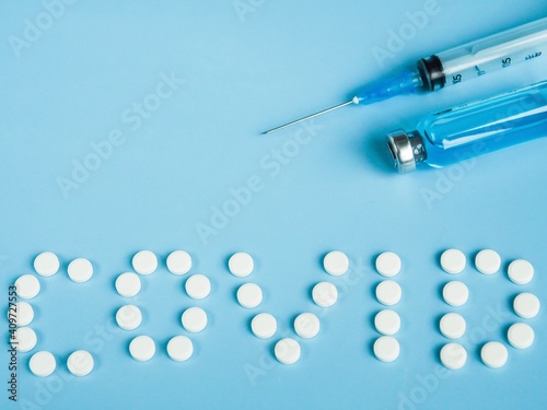 The inscription Covid , made of white round tablets. Disposable syringe and vial drug on a blue background. COVID-19 coronavirus vaccine. Space for text