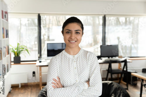 Confident Indian millennial businesswoman posing with hands folded in office. Head shot portrait of happy young female professional, business leader, boss or corporate coach looking at camera.