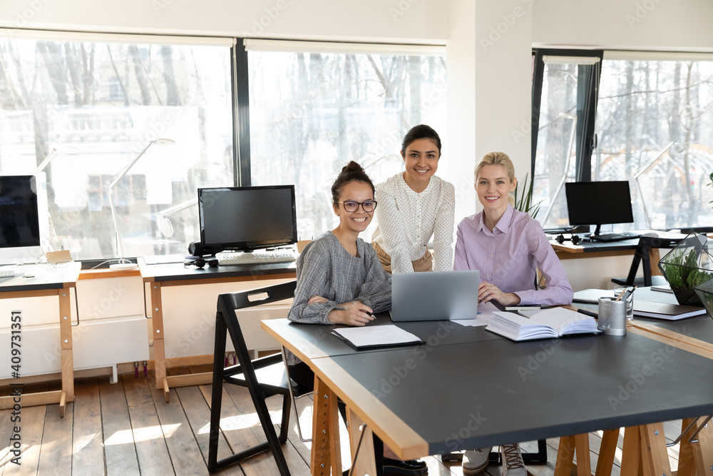 Portrait of mixed race employees and female business team leader at office workplace. Indian coach and interns posing during training at laptop, looking at camera, smiling. Diverse professional group