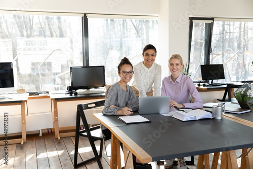 Portrait of mixed race employees and female business team leader at office workplace. Indian coach and interns posing during training at laptop, looking at camera, smiling. Diverse professional group