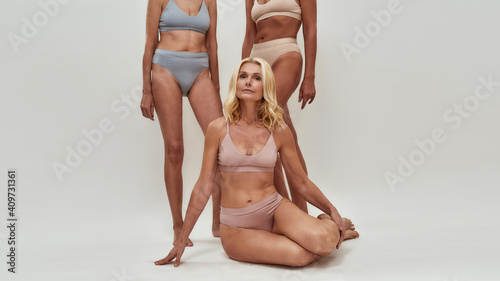 Attractive caucasian senior woman in underwear looking at camera while sitting half naked in studio isolated over light background. Women standing in the background