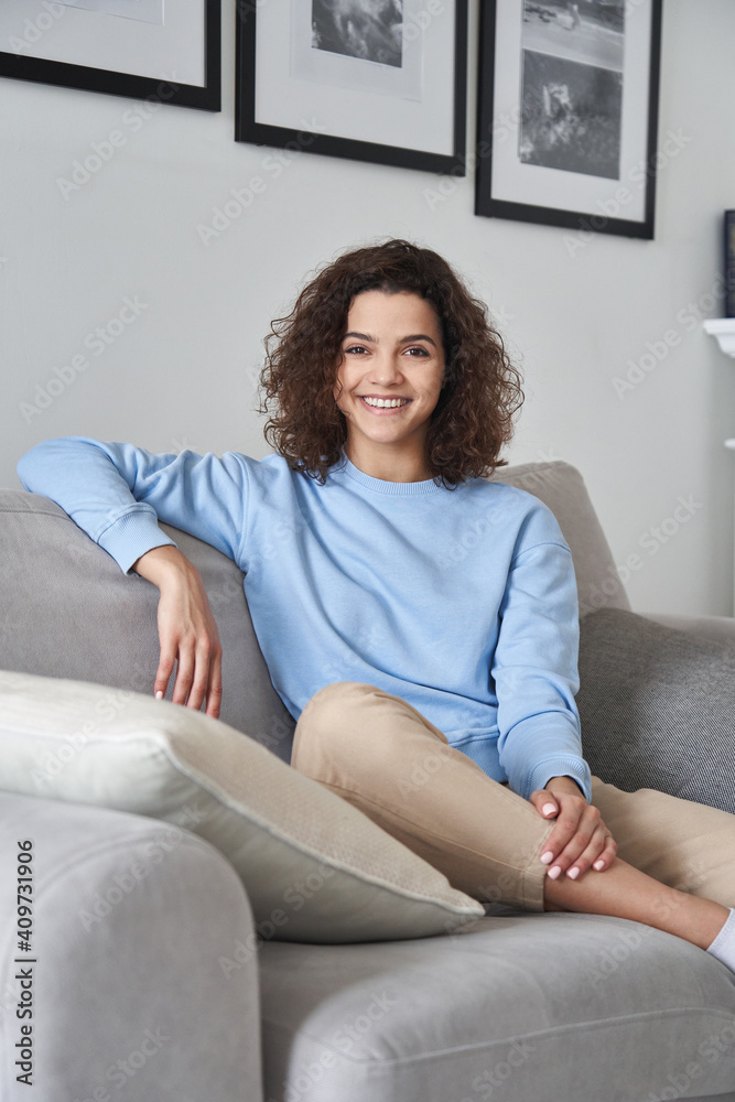 Smiling young pretty hispanic woman looking at camera sitting on comfortable sofa at home. Happy cheerful casual teen girl, female model posing indoors, relaxing on couch in modern apartment, portrait Smiling young pretty hispanic woman looking at camera sitting on comfortable sofa at home. Happy cheerful casual teen girl, female model posing indoors, relaxing on couch in modern apartment, portrait