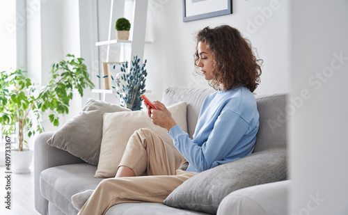Hispanic teen girl holding cell phone sitting on sofa at home. Young latin woman using smartphone tech mobile apps online services on cellphone device relaxing in apartment living room.