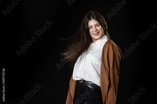 chica joven moviendo el pelo y posando con chaqueta marron y fondo negro