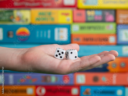 Hand with white dice on the blurred background of colorful board game boxes. Concept of entertainment during pandemic of covid-19