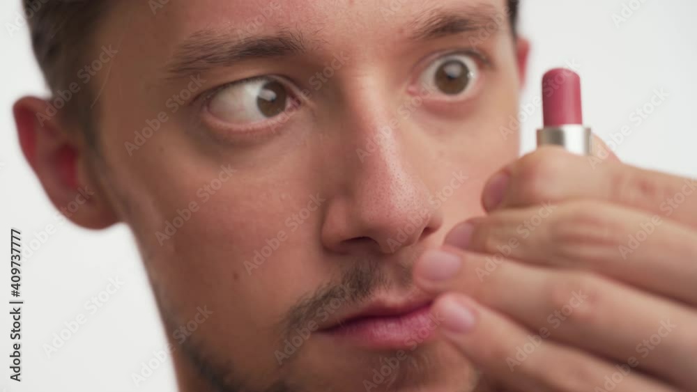 Close up portrait of one young stupid guy with brown eyes, hair, beard stare at pink lipstick, play with it, open and close on white background. Dumb male cross eyed, focus on women's accessory indoor