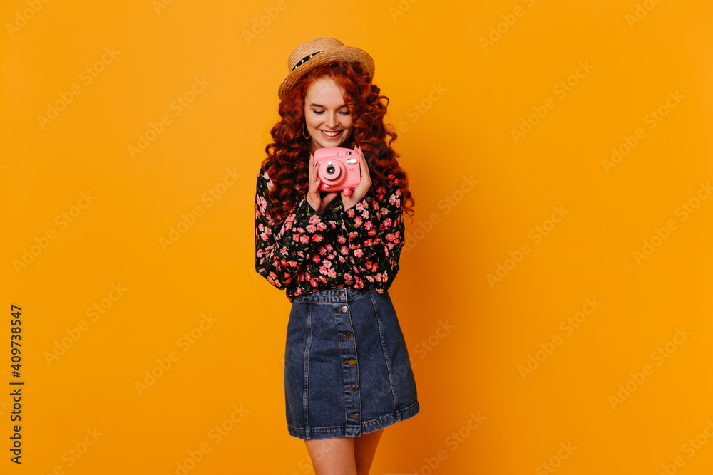 Charming woman with smile is holding pink mini camera. Lady in stylish denim outfit and straw hat posing in orange studio Charming woman with smile is holding pink mini camera. Lady in stylish denim outfit and straw hat posing in orange studio