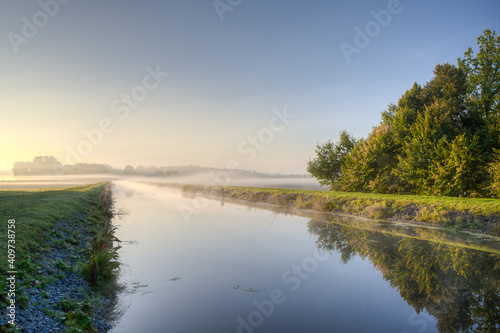 A autumn morning at the Schaalsee Canal, fog drifts over the smooth surface of the water and the rising sun bathes the landscape in its warm yellow-orange colored light..