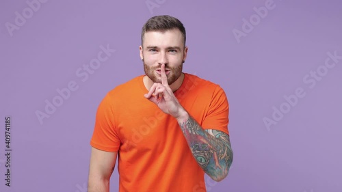 Secret bearded tattooed young man 20s in orange basic t-shirt isolated on purple violet background studio. People lifestyle concept. Looking aside saying hush be quiet with finger on lips shhh gesture