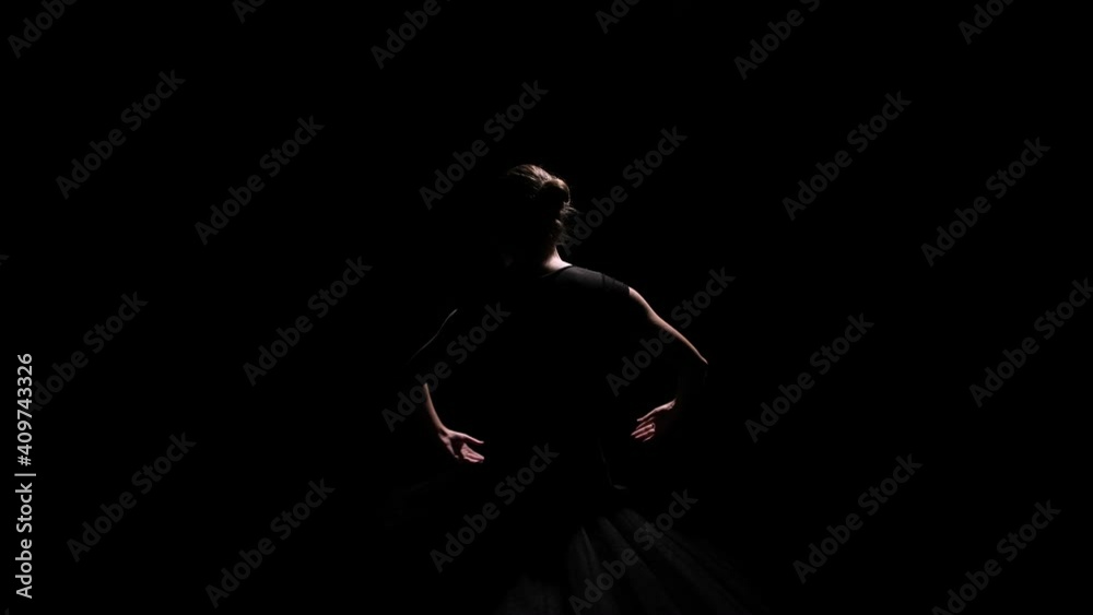 Camera rotates around young flexible ballerina in black tutu. Feminine female gracefully moves her arms while performing against a black studio background. Silhouette. Close up. Slow motion.