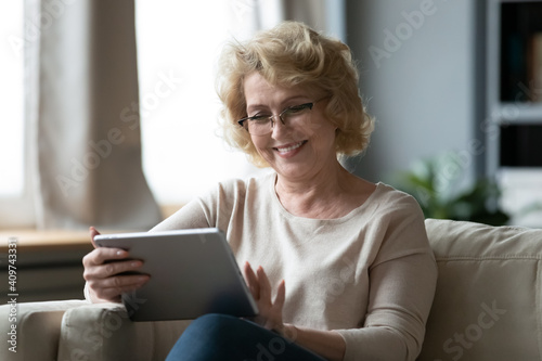 Happy middle aged older retired woman in eyeglasses using modern digital computer tablet, relaxing on sofa. Smiling senior granny enjoying web surfing or shopping online, easy technology usage.