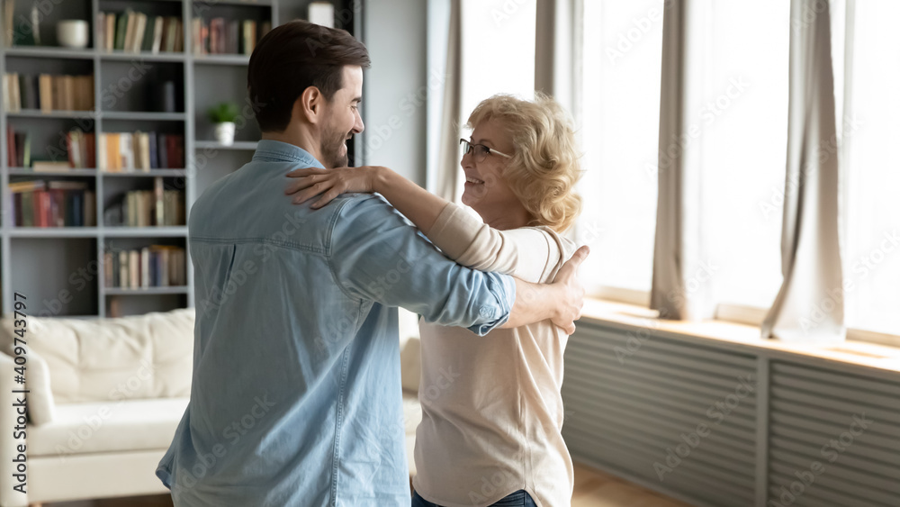 Fototapeta premium Smiling young caucasian man dancing with happy older middle aged beautiful mother in living room, enjoying weekend family hobby activity at home, grown children and retired parents pastime concept.