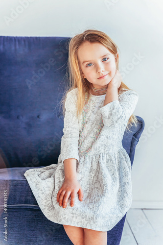 Sweet little girl in white dress sitting on modern cozy blue chair relaxing in white bright living room at home indoors. Childhood schoolchildren youth relax concept