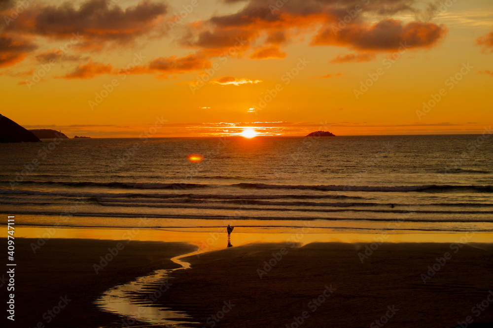 Surfer looking out to sea at sunset in Cornwall. Beach with surfer holding surfboard in Summer. Cornish beach surfer. Surfer looking out to sea at sunset in Cornwall. Beach with surfer holding surfboard in Summer. Cornish beach surfer.