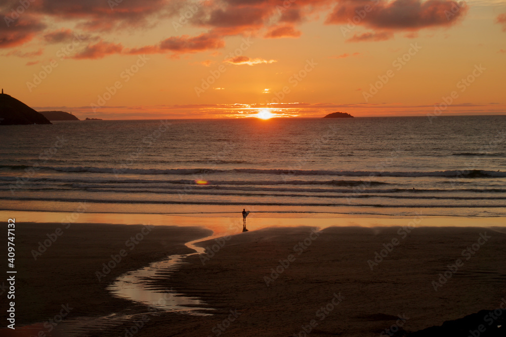 Surfer looking out to sea at sunset in Cornwall. Beach with surfer holding surfboard in Summer. Cornish beach surfer. 