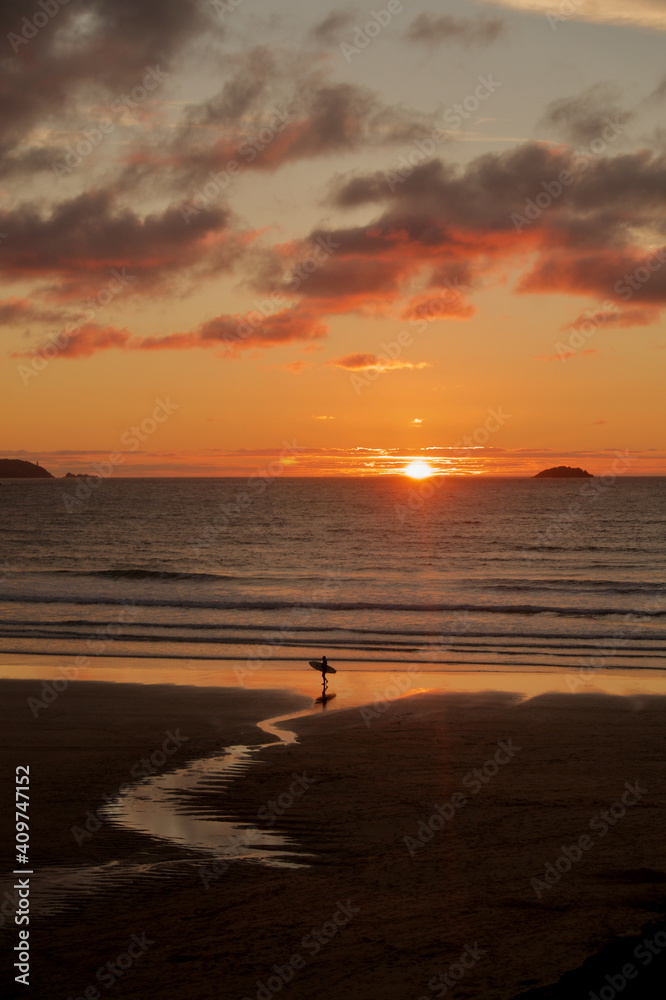 Surfer looking out to sea at sunset in Cornwall. Beach with surfer holding surfboard in Summer. Cornish beach surfer. 