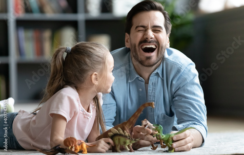 Overjoyed young father laughing, joking with happy small adorable child daughter while playing toys together on floor carpet in living room, different generations family entertaining activity concept.