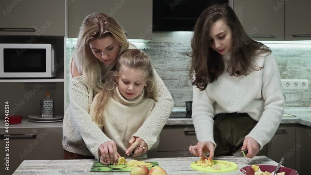 Young mother and two her daughters cooks fruits salad at kitchen. Mother teaches her little daughter to cut orange Young mother and two her daughters cooks fruits salad at kitchen. Mother teaches her little daughter to cut orange