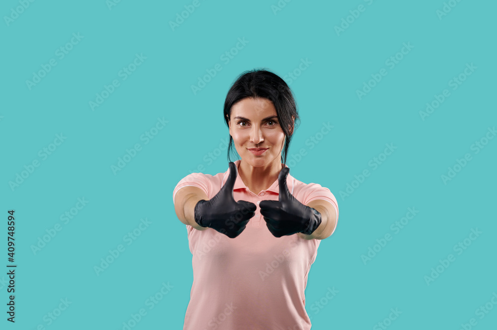 Smiling beautiful woman wearing black protective latex gloves showing thumbs up. Portrait isolated on blue background