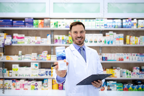 Shot of handsome male pharmacist standing at the drugstore counter and holding vitamins for sale. In background shelf with medicines.