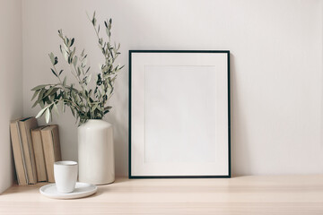 Breakfast still life. Cup of coffee, books and empty picture frame mockup on wooden desk, table. Vase with olive branches. Elegant working space, home office concept. Scandinavian interior design.