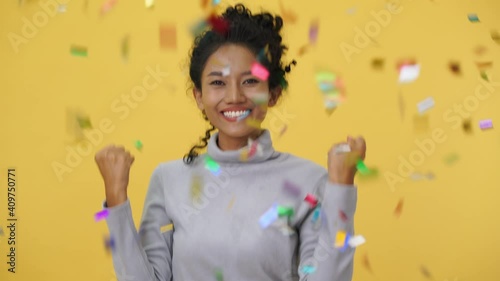 Celebrating young woman excited girl rejoices over confetti rain isolated over yellow orange background in studio