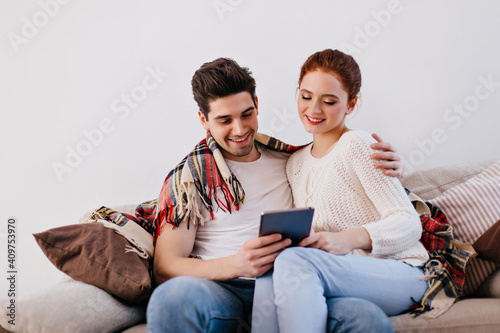 Smiling young people sitting on couch in living room. Man embracing wife while using tablet.