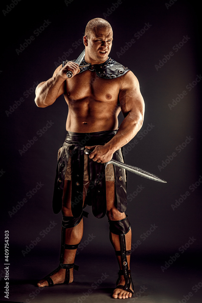 Severe barbarian in leather costume with two swords. Portrait of balded muscular gladiator. Studio shot. Black background.