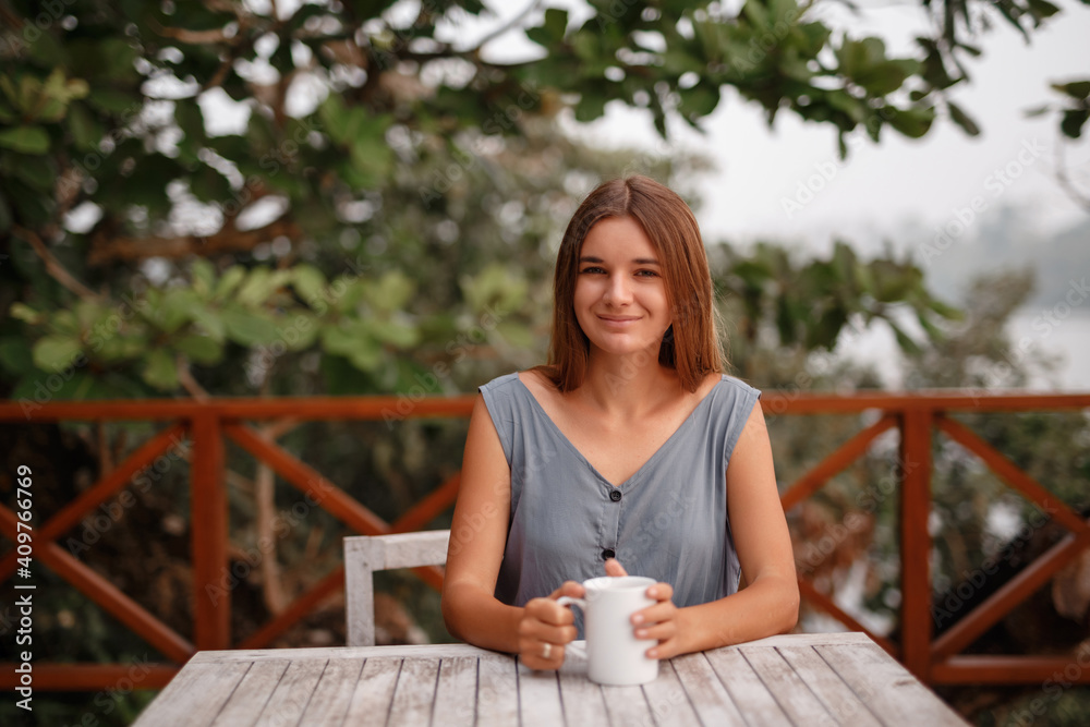 Portrait of young beautiful girl, holding a warm cup with tea or coffee on the background of nature near the river, look at the camera Portrait of young beautiful girl, holding a warm cup with tea or coffee on the background of nature near the river, look at the camera