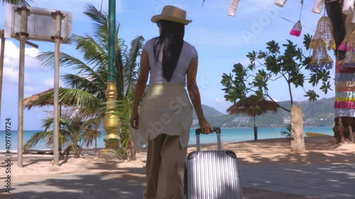 4K Asian travel women walking with her travel bag on side road at the beach during a sunny day view. Shot from behind. Happy women tourists walk with suitcases on the beach road. Travel holiday summer