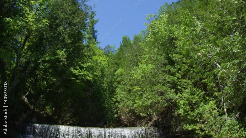 Camera tilts down to reveal a young woman in a bikini standing in a river, waterfall in background. Wide shot, push in. Camera tilts down to reveal a young woman in a bikini standing in a river, waterfall in background. Wide shot, push in.