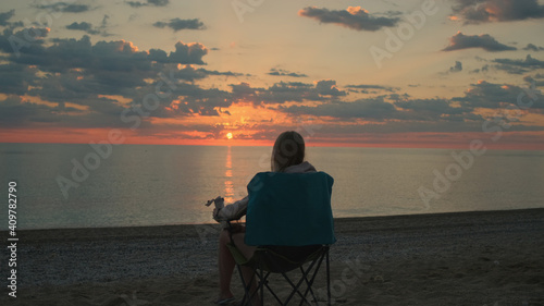 Medium shot of woman sitting on camping chair and drinking coffee. Relaxing and enjoying nature. Admiring sunrise on the sea.