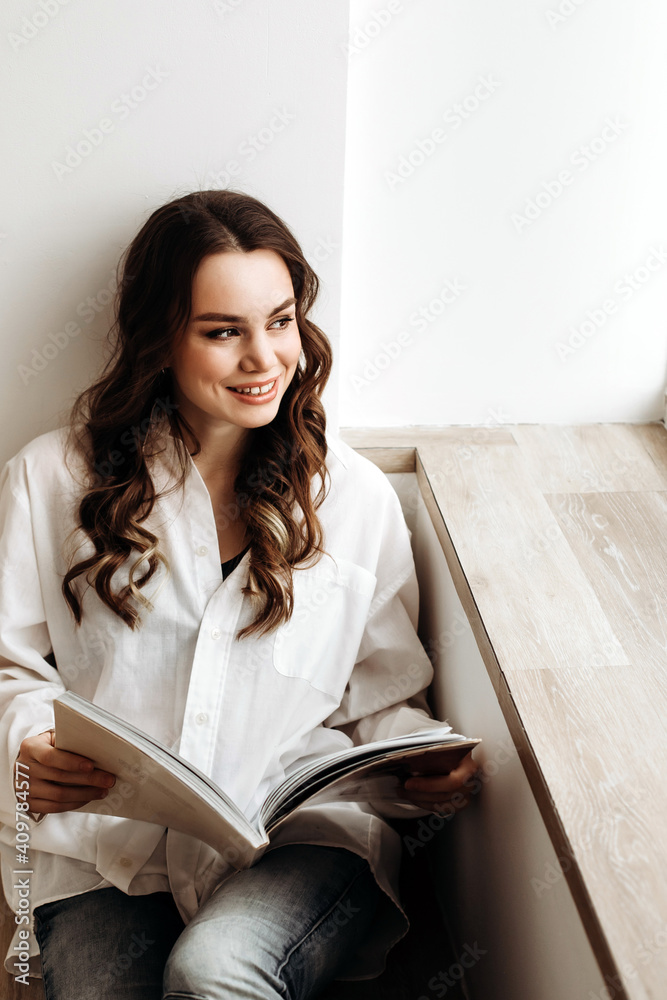 woman of European appearance reads a book. Portrait of a lady in a white shirt with a magazine in her hand. Woman sitting indoors by the window on the windowsill woman of European appearance reads a book. Portrait of a lady in a white shirt with a magazine in her hand. Woman sitting indoors by the window on the windowsill