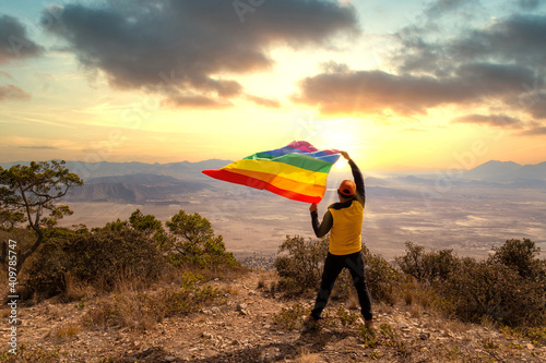 Back view of a man standing on top of the hill and holding the LGBT pride flag