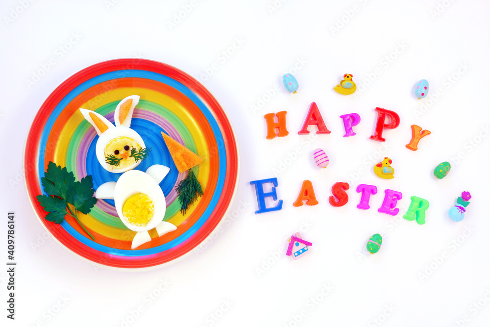 Easter breakfast idea for kids. Bunny rabbit made of boiled eggs on multicolored plate, white background, inscription Happy Easter, top view 