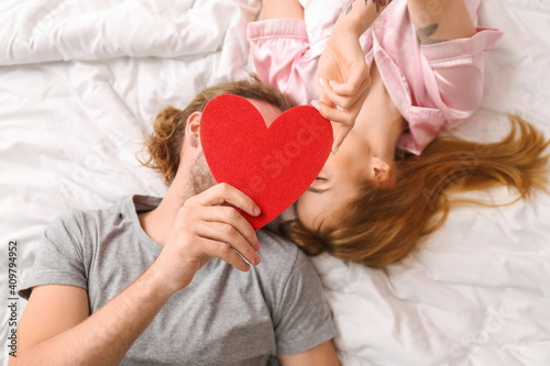 Young couple with red heart lying on bed, top view. Valentine's Day celebration