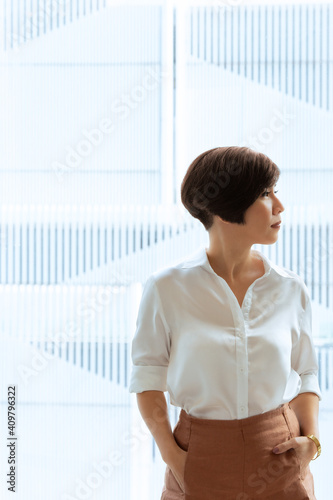 Free Download Portrait of beautiful, confident , short hair and smart looking Asian businesswoman standing by window in office, putting hands in pocket, looking out with modern skyscraper in background. - GFXLoad