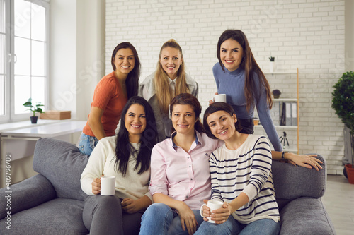 Group of young women sitting on sofa at home, drinking tea, smiling and looking at camera. Happy friends and coworkers enjoying Casual Friday at work and spending coffee break on modern office couch