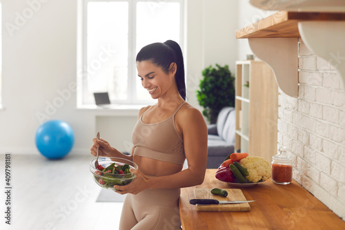 Slender fitness woman in sportswear standing at home in the kitchen with a bowl of fresh salad. Happy woman enjoys healthy food after workout. Fitness, dieting and healthy lifestyle concept.