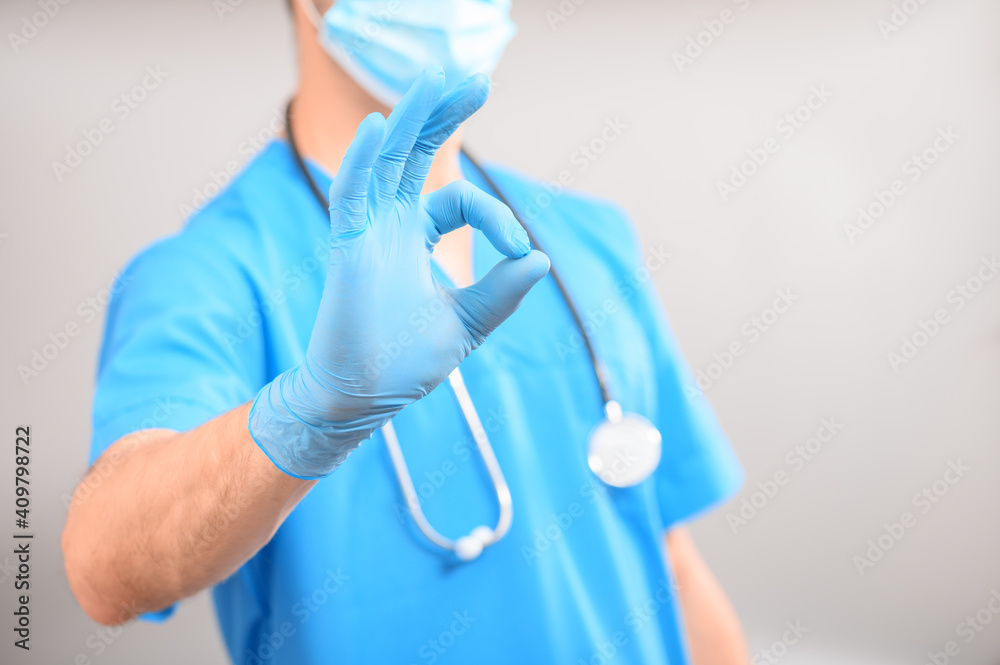 A doctor in a blue uniform wearing a medical mask with a stethoscope around his neck shows the Ok gesture, against a uniform gray background