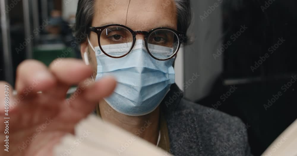 Close up view of smart man in medical mask turning book page while sitting in bus. Male person in wireless earbuds reading story in public transport in evening. Concept of commuting. Close up view of smart man in medical mask turning book page while sitting in bus. Male person in wireless earbuds reading story in public transport in evening. Concept of commuting.