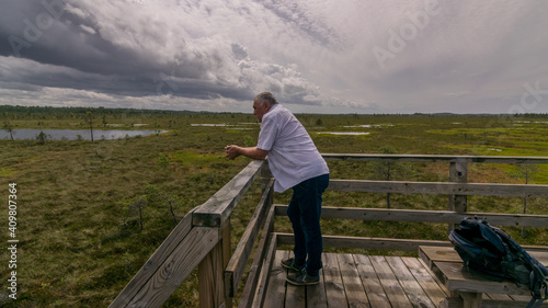the summer swamp. a man in a white shirt looks out of a wooden tower. bog background and vegetation. white clouds. small swamp pines