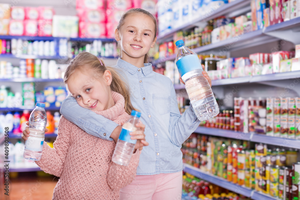 Portrait of cheerful little girls standing among shelves in the supermarket, holding a bottled water