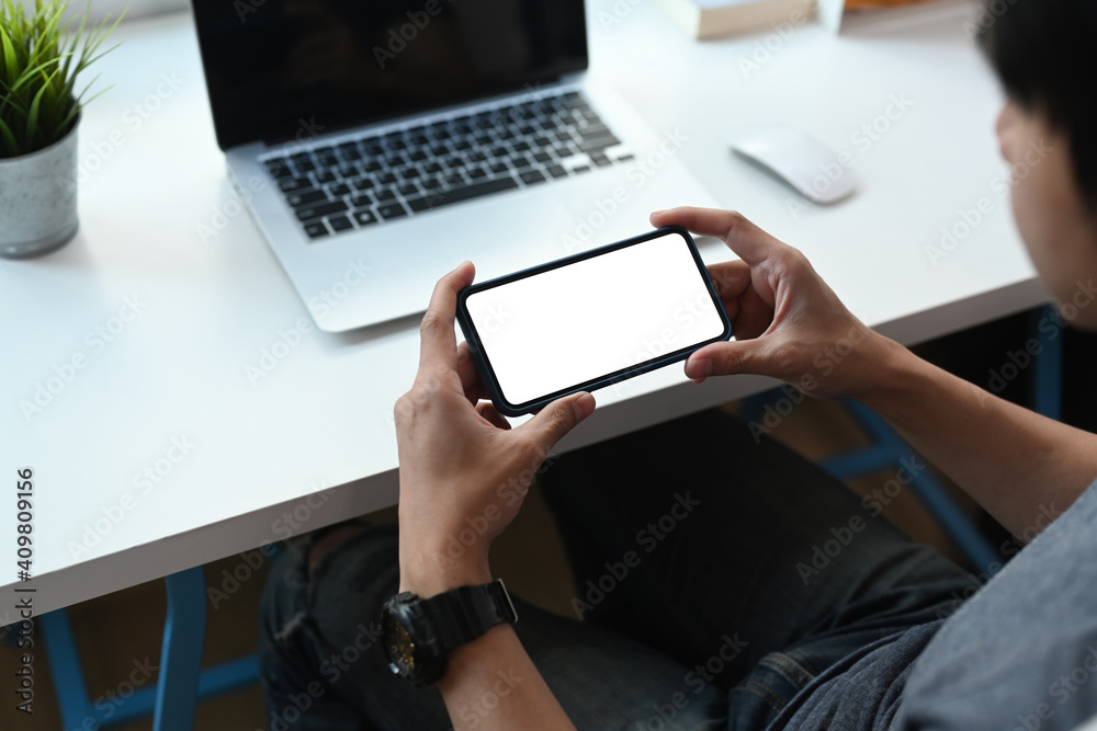 Young man sitting at his workspace and holding horizontal mobile phone checking social network. Young man sitting at his workspace and holding horizontal mobile phone checking social network.