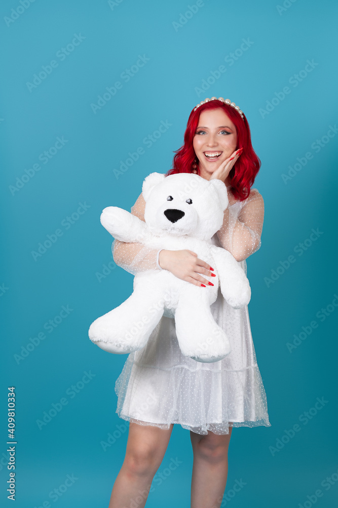 surprised delighted young woman a woman with an open mouth, with red wavy hair in a white dress hugs a large white teddy bear, isolated on a blue background. surprised delighted young woman a woman with an open mouth, with red wavy hair in a white dress hugs a large white teddy bear, isolated on a blue background.