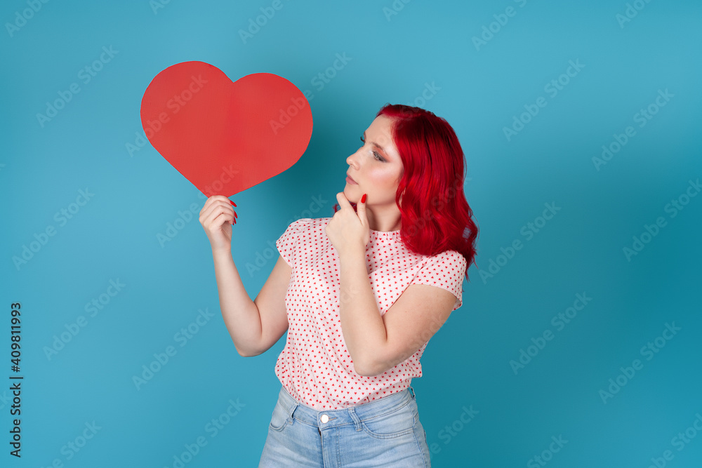 close-up pensive dreamy young woman with red hair holds a large red paper heart and looks at it isolated on a blue background close-up pensive dreamy young woman with red hair holds a large red paper heart and looks at it isolated on a blue background