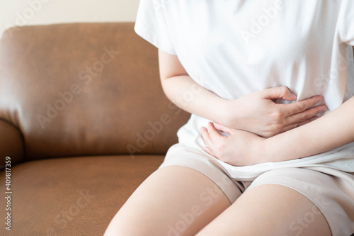 Young female suffering form stomach ache while sitting on couch at home. Causes of abdominal pain include menstruation pain, gastritis, stomach ulcer, food poisoning, diarrhea or IBS.