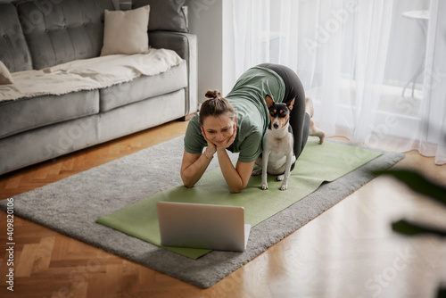 Cheerful beautiful woman practicing yoga at home online from laptops by video chatting on the carpet and yoga mate with her dog nearby, sports and mental health