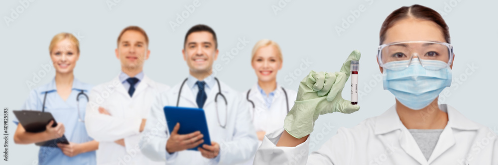 medicine, health and pandemic concept - young asian female doctor or scientist in medical mask and goggles holding beaker with blood test over medical team on blue background medicine, health and pandemic concept - young asian female doctor or scientist in medical mask and goggles holding beaker with blood test over medical team on blue background