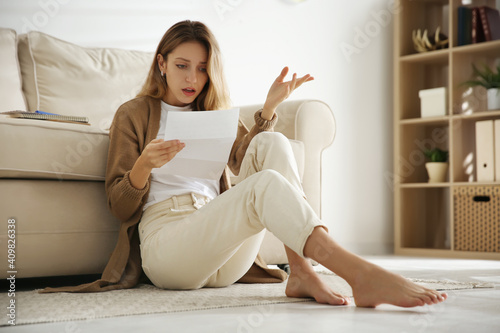 Shocked woman reading letter while sitting on floor near sofa at home photo